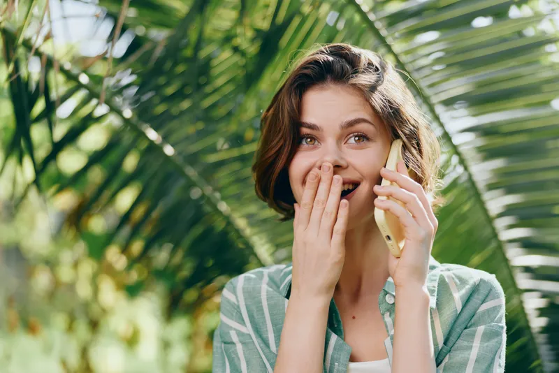 woman outdoors smiling while talking on her mobile phone, casual wear, tropical garden backdrop, sunny day, candid lifestyle portrait, cheerful mood, natural light, relaxed pose, friendly