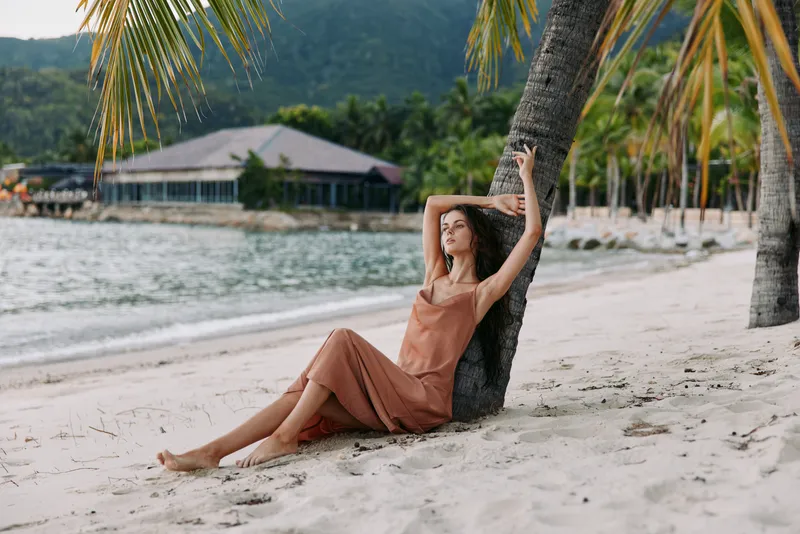 A serene portrait on a sandy tropical beach shows a woman resting against a leaning palm tree, wearing an airy orange dress as gentle waves lap the shore.