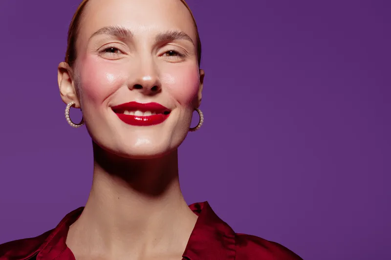 Smiling woman with red lipstick and hoop earrings poses against purple background. Close-up portrait with smooth skin and confident expression. Concept of beauty and makeup.
