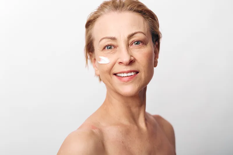 Smiling mature woman with natural skin applies moisturizing cream on cheek. Close-up portrait on white background. Concept of skincare, aging beauty, wellness, and self-care routine.