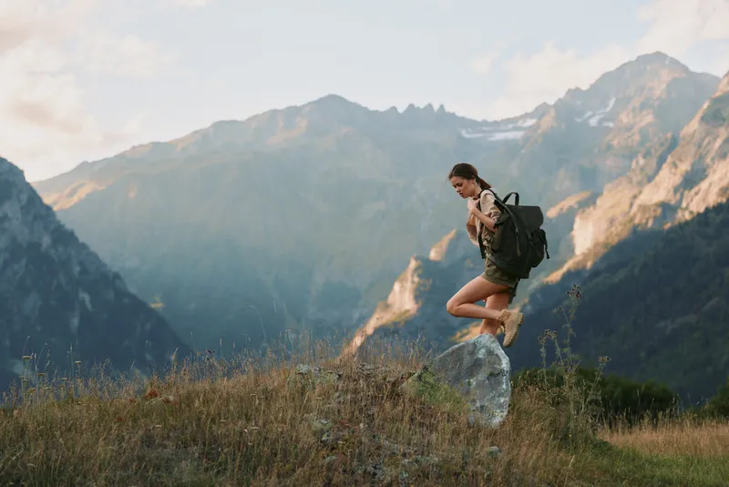 A woman hiking in outdoor mountains jumps over grass, wearing a backpack and casual attire. This scene conveys adventure, fitness, active lifestyle, nature, landscape.