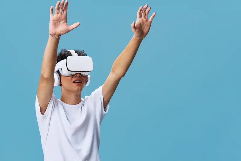 young man wearing white t-shirt using virtual reality headset with raised hands on blue background, immersive technology experience, casual indoors