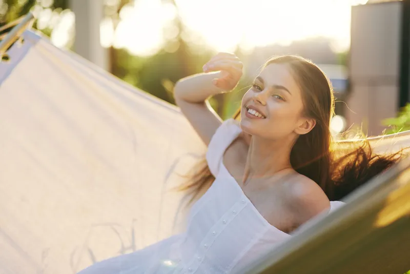 Young woman relaxing in a hammock at sunset, wearing a white summer dress, radiating joy and tranquility, perfect for summer lifestyle concepts