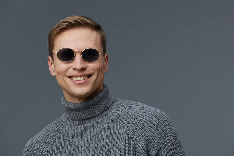 Smiling man wearing black sunglasses and gray turtleneck sweater look at camera pose studio portrait on plain gray background. People lifestyle concept
