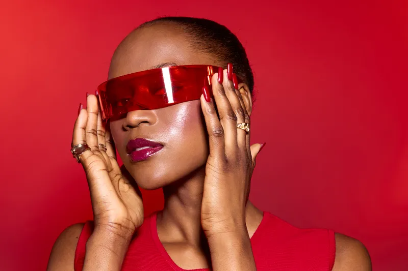 Portrait of a dark skinned African woman wearing red futuristic sunglasses and red top, posing with hands near face on red background. Concept of fashion, beauty, and modern style.