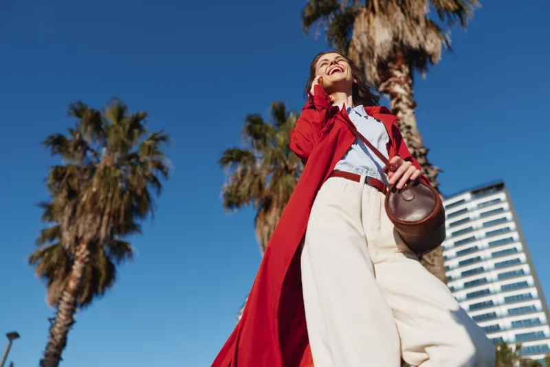 A stylish woman in a red trench coat strolls outdoors under a bright blue sky, carrying a round leather bag and smiling confidently.