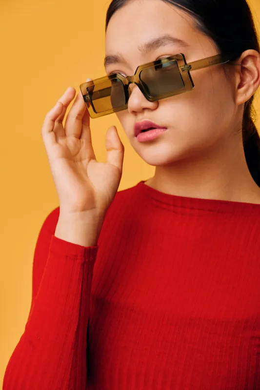 Young woman wearing oversized geometric sunglasses and a red sweater posing against a yellow background. Fashion portrait with trendy eyewear and casual style closeup.