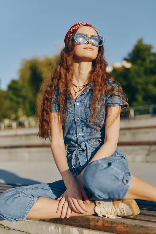 Lifestyle woman in modern boho-western denim street style sitting outdoors wearing sunglasses and a red headband captured in warm film color tones.