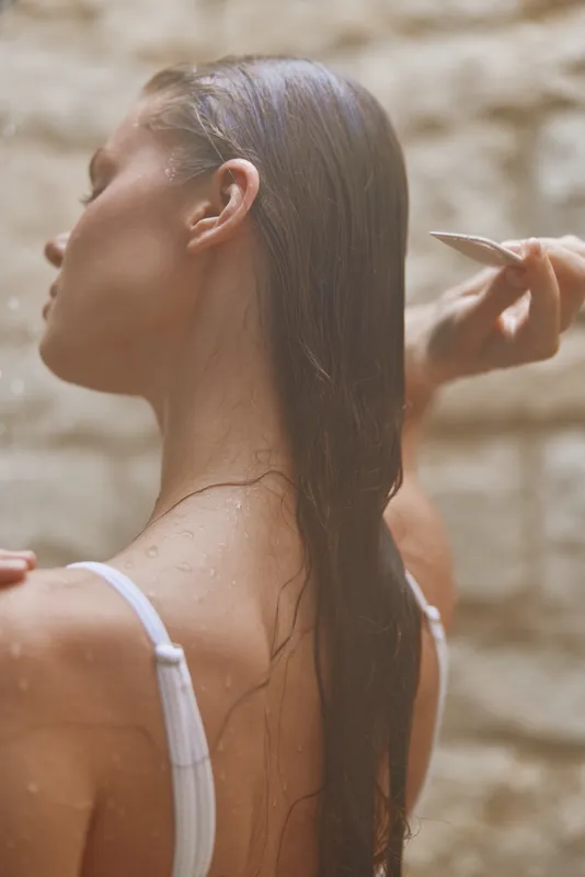 Woman combing wet long hair under natural light with blurred stone wall background, showing casual morning routine and personal care moment.