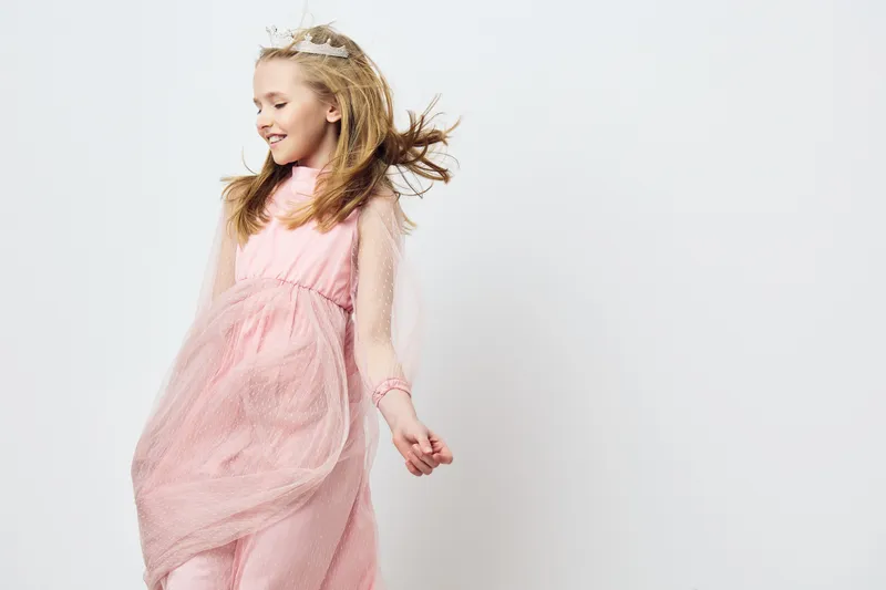 Playful girl in a pink dress with flowing fabric, joyfully dancing against a soft white background, showcasing innocence and happiness in childhood