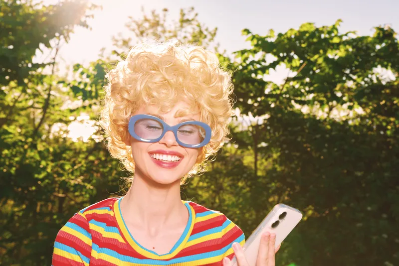 A cheerful woman in a colorful striped shirt and retro blue glasses uses a smartphone outdoors, bright park light emphasizing playful lifestyle and retro charm.
