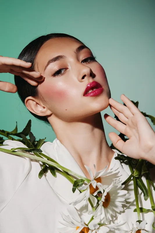 Young woman with glossy red lips and highlighted cheeks poses with green foliage and white flowers on a bright background. Beauty fashion creative portrait concept.
