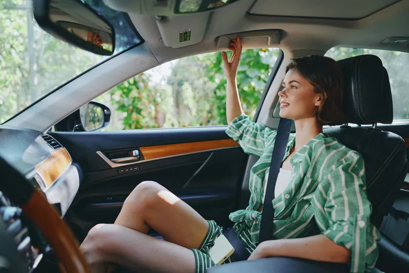 A relaxed woman sits in a car’s front passenger seat, wearing a seatbelt and a casual green striped shirt, ready for a drive.