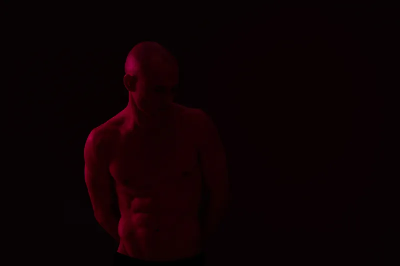 A contemplative man with a toned physique, posed against a dramatic dark background, illuminated by soft red light, creating an evocative, intimate atmosphere.