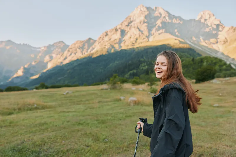 A woman hiking in a scenic mountain landscape. She smiles softly, enjoying the outdoors and fresh air, captured in warm golden hour light.