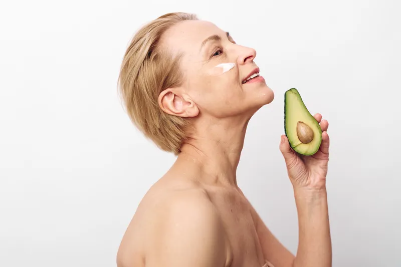 Mature woman with short blonde hair holds fresh avocado near face, smiling with natural skin cream on cheeks. Concept of healthy skincare, nutrition, beauty, and self-care for aging skin.