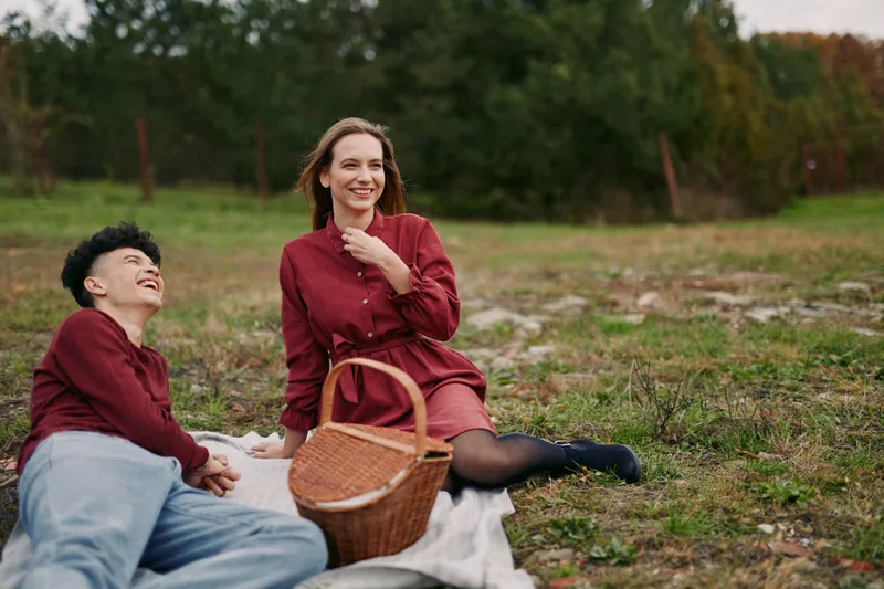 Authentic moment captures two companions outdoors in a quiet field, relaxed pose, candid smiles, warm light, simple picnic, genuine connection under open sky.