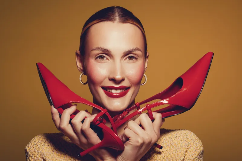 Smiling woman with red lipstick holds red high heel shoes near face on yellow background. Concept of fashion, style, beauty, and confident female model with elegant accessories.