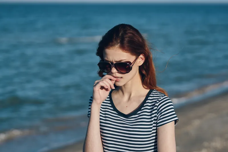 Женщина в солнечных очках стоит на пляже у моря, на заднем плане — природный пейзаж