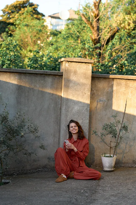 Cheerful woman lifestyle portrait, seated on ground in a red dress, enjoying a sunlit garden moment, relaxed and natural, against a weathered stone wall