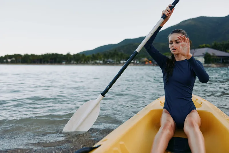 Young paddler sits in a yellow kayak, raising an oar on calm coastal water, wearing a navy swimsuit, with distant hills and clear sky framing an energetic outdoor scene.