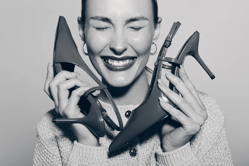 Happy young Caucasian woman with fair skin smiles with closed eyes holding two high heel shoes near face in cozy knitted sweater. Concept of fashion and joy.