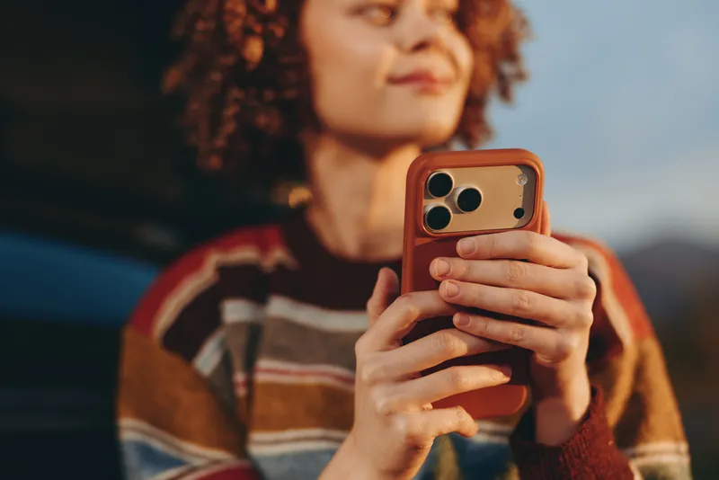 Woman lifestyle outdoors holding smartphone with both hands, curly hair framing her face, warm knit sweater, calm expression, soft daylight, blurred background, relaxed candid moment
