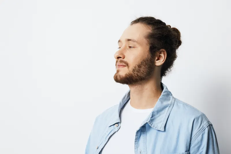 Casual portrait of a young man with curly hair and a beard, wearing a denim jacket, exuding confidence against a light background