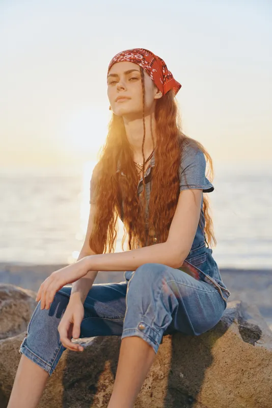 lifestyle woman in modern boho-western denim street style with red bandana, long hair, and relaxed pose in warm film color near the ocean at sunset