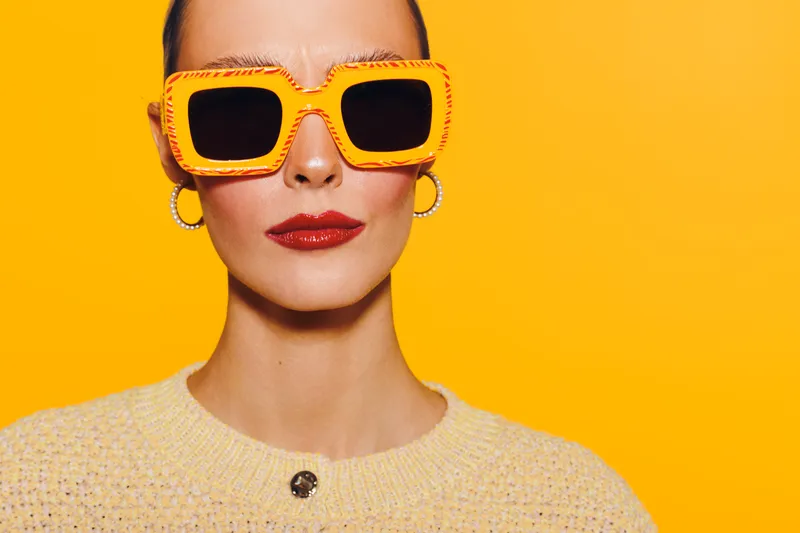 Stylish woman with bright orange sunglasses and red lipstick poses against vibrant yellow background. Fashion portrait with modern accessories and confident expression.
