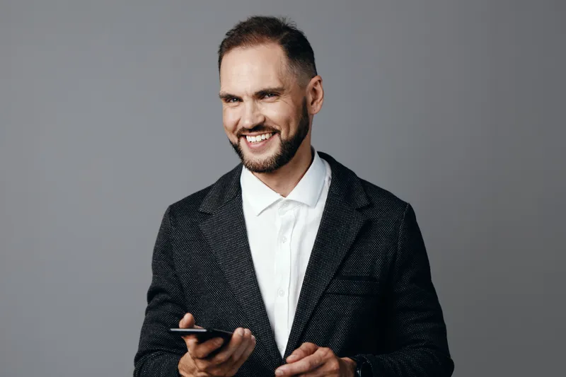 A professional man in business attire uses a handheld device, smiling confidently against an isolated colored background, conveying focus, warmth, and poised energy.