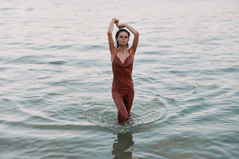 A woman stands waist deep in calm water, wearing a red jumpsuit, arms lifted overhead, creating a serene, cinematic mood with soft reflections and graceful, effortless posture.