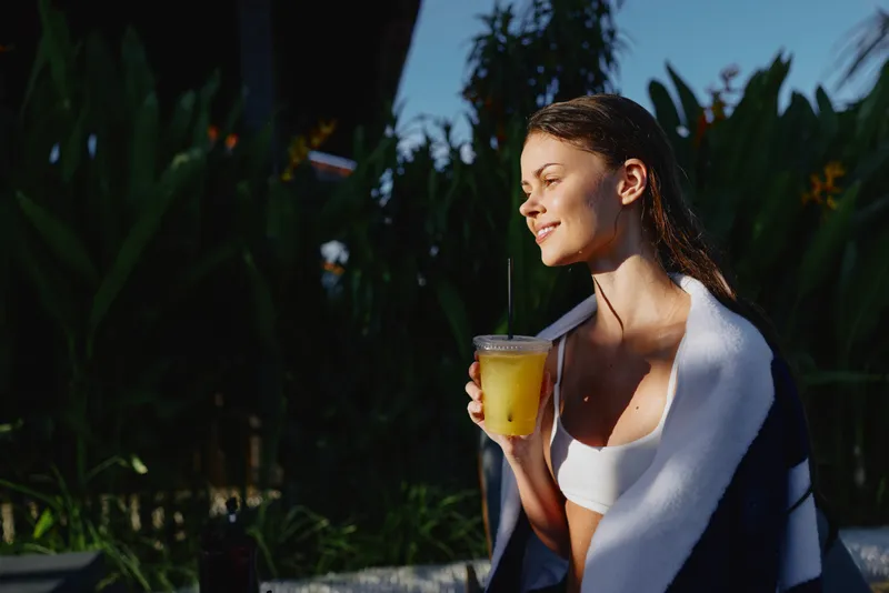 Smiling woman with towel on her shoulders sipping orange juice outdoors, poolside backdrop, relaxed lifestyle, wellness and selfcare, warm natural light creating a sunny, inviting mood