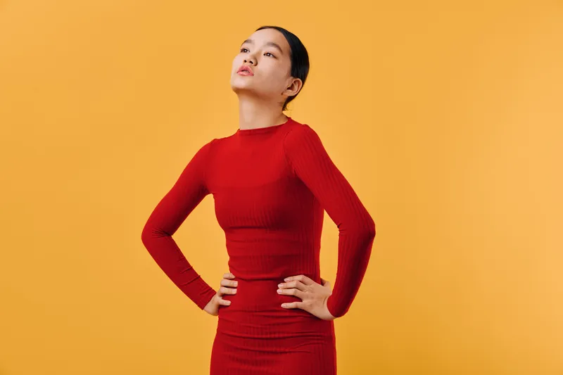 Young woman in red dress stands with hands on hips and confident posture against yellow background, posing with calm expression and elegant style for fashion portrait.