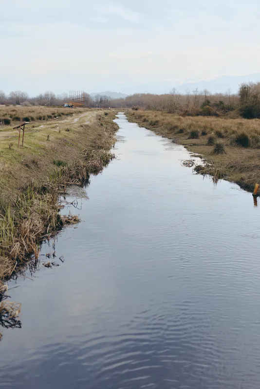 Calm river winding through rural meadow and grassy banks, soft ripples on water, leafless trees and cloudy sky in distance, outdoor nature landscape lifestyle travel concept