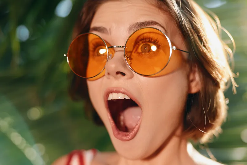 A cheerful young woman wearing round orange sunglasses is surprised and excited, captured in bright natural light. Her open mouth and wide eyes convey playful astonishment.