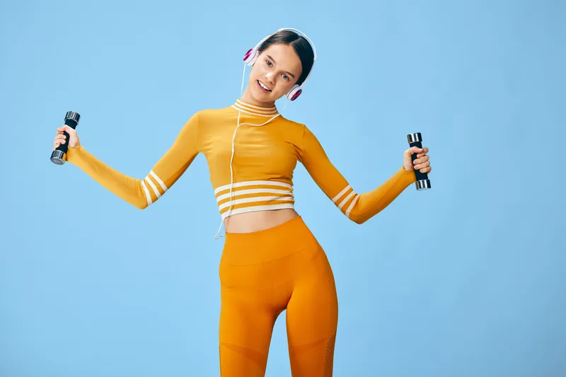 Woman fitness scene: young woman in orange activewear holding dumbbells and wearing headphones, smiling and exercising with light arm lifts against a clean blue studio background.