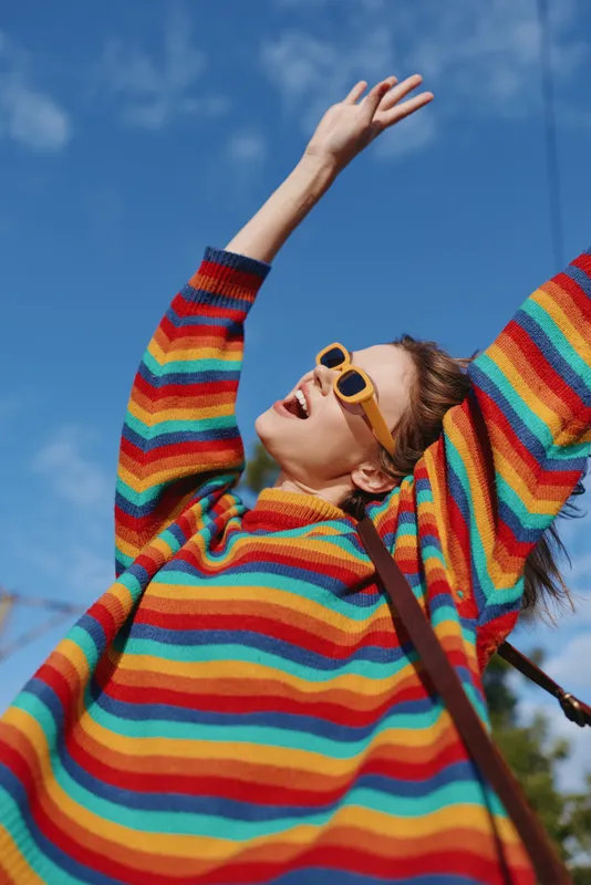 Woman in her mid 20s wearing a bright rainbow sweater and yellow sunglasses, smiling with arms raised under clear blue sky in an urban park, joyful travel lifestyle casual outfit.