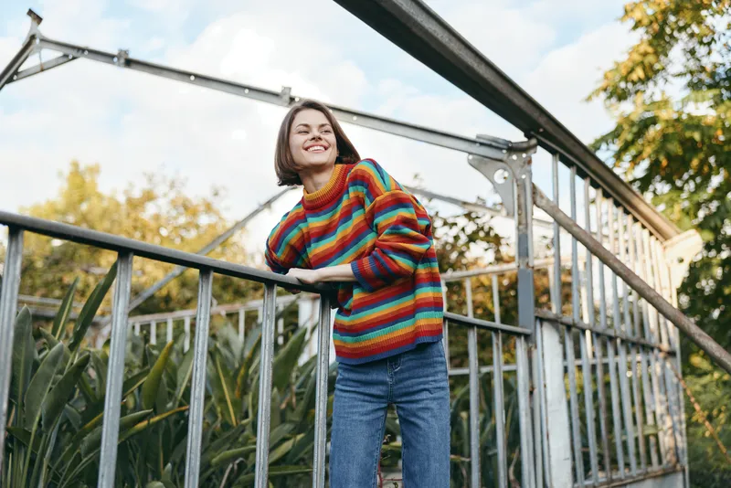 Woman in colorful striped sweater smiling and leaning on a metal railing at a greenhouse urban garden. Young adult in casual jeans with relaxed joyful expression for lifestyle fashion.