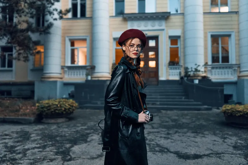 Модная женщина в красном берете и кожаной куртке принимает эффектную позу на фоне городского здания в солнечный день