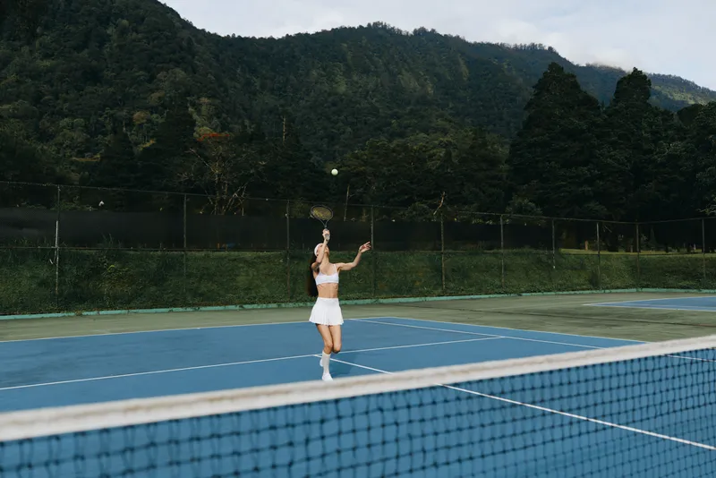 Young female tennis player in white sportswear, serving on outdoor court surrounded by lush green mountains, showcasing determination and athleticism