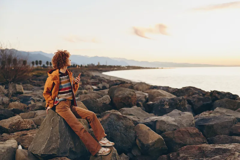 Woman lifestyle, sitting on rocky beach at sunset, travel mood on rocks, curly hair, casual outfit, gazing at calm sea, distant mountains, warm golden light, serene mood