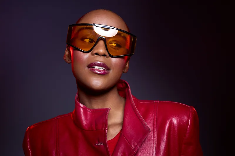 Confident black woman with dark skin wearing oversized orange sunglasses and a red leather jacket poses against dark background. Fashion portrait with bold style and modern accessories.