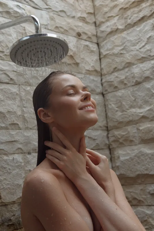 Relaxed woman under shower with eyes closed enjoying water flow, natural stone background, calm expression, wellness and self-care moment indoors.