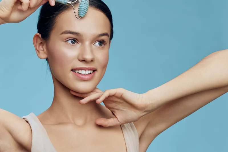 A smiling young woman using a facial roller for skincare, showcasing natural beauty against a soft blue background, emphasizing self care and wellness in a modern aesthetic