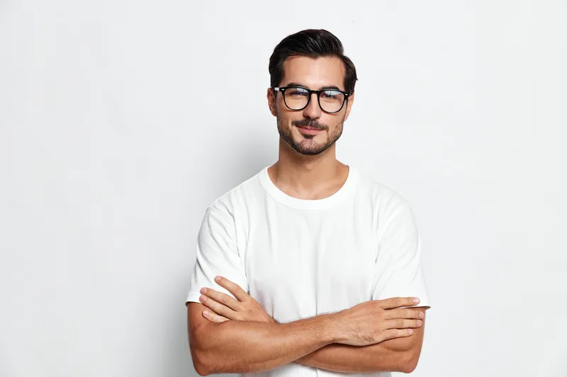 Smiling man with glasses wearing a white t-shirt, standing confidently with arms crossed against a plain solid background. Portrait for lifestyle concept.