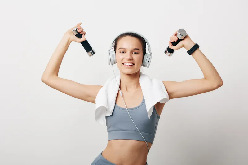 Female athlete in athletic wear lifts dumbbells with a towel over her shoulders, wearing over-ear headphones, exuding energy during a focused fitness routine in a clean studio.