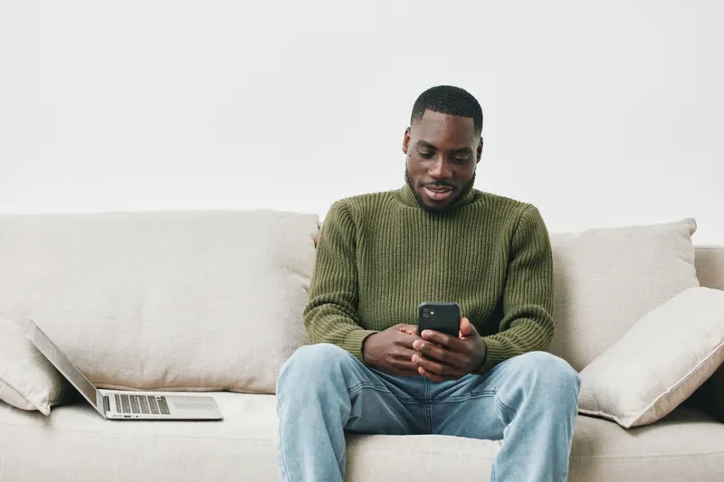 Young Black man sitting on a sofa, smiling while using a smartphone, wearing a green sweater and jeans in a modern, bright living space