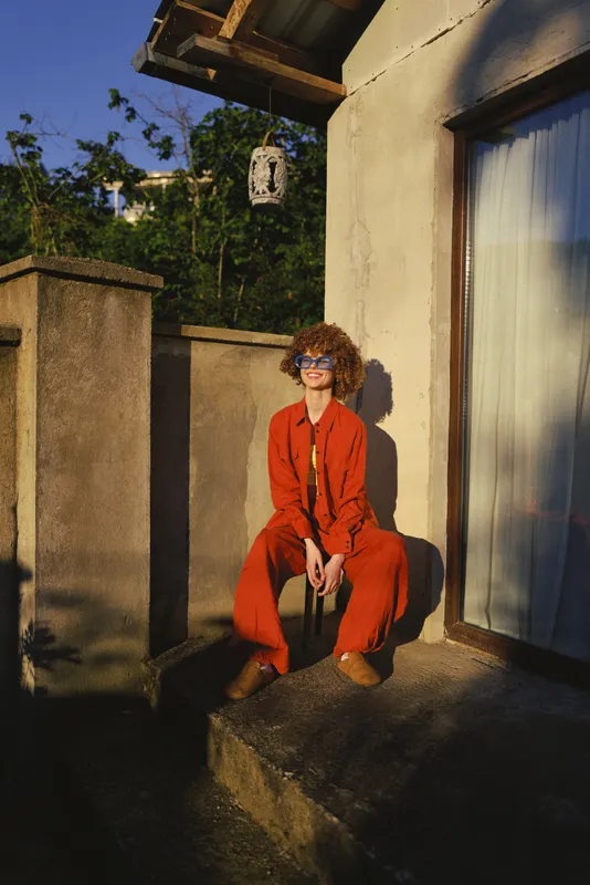 Vibrant woman outdoors, smiling in sunlight, red coat, relaxed pose on a sunny terrace, natural colors and warm tones, casual lifestyle moment, soft breeze, natural charm flair