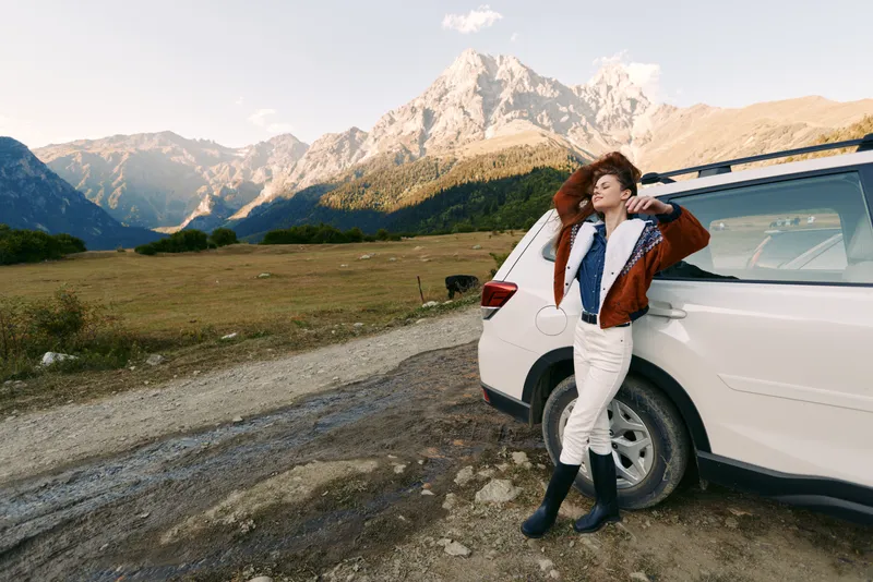 Woman leaning against white car SUV on roadside in mountains nature landscape, enjoying travel and road journey with relaxed pose, jacket and boots, scenic countryside horizon.
